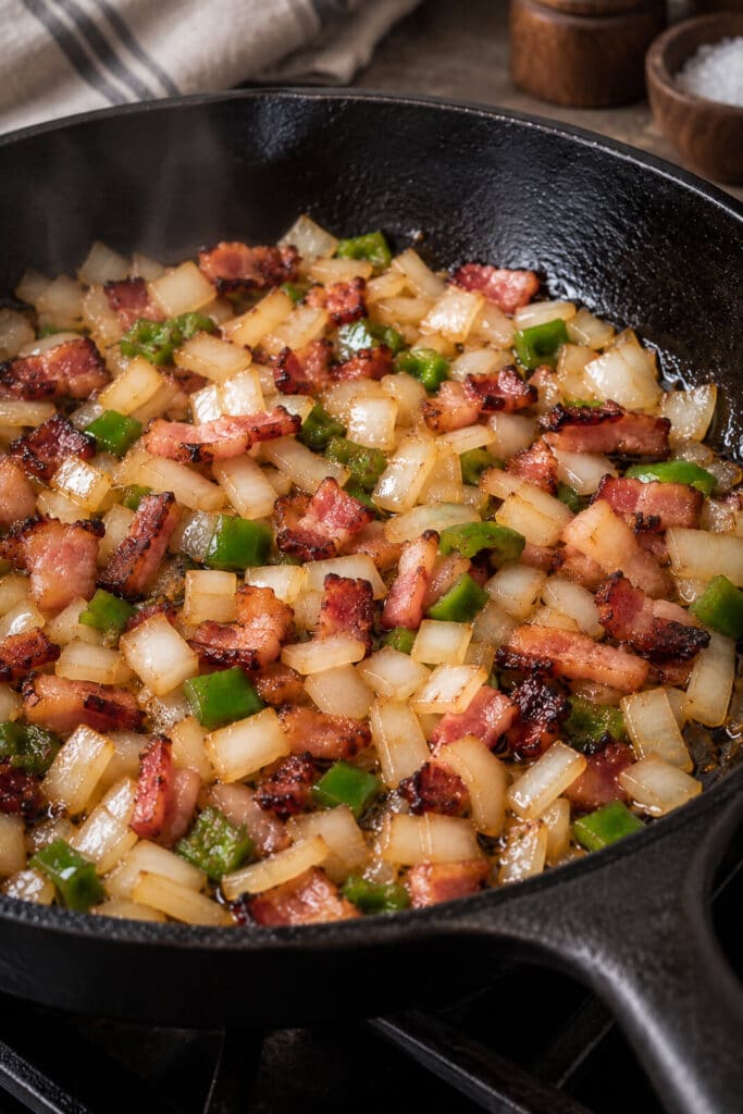 bacon, onions, and peppers cooking down in a cast iron pan