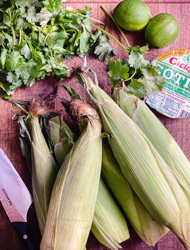 corn on the cob still in it's husks with cojita cheese and limes on a cutting board