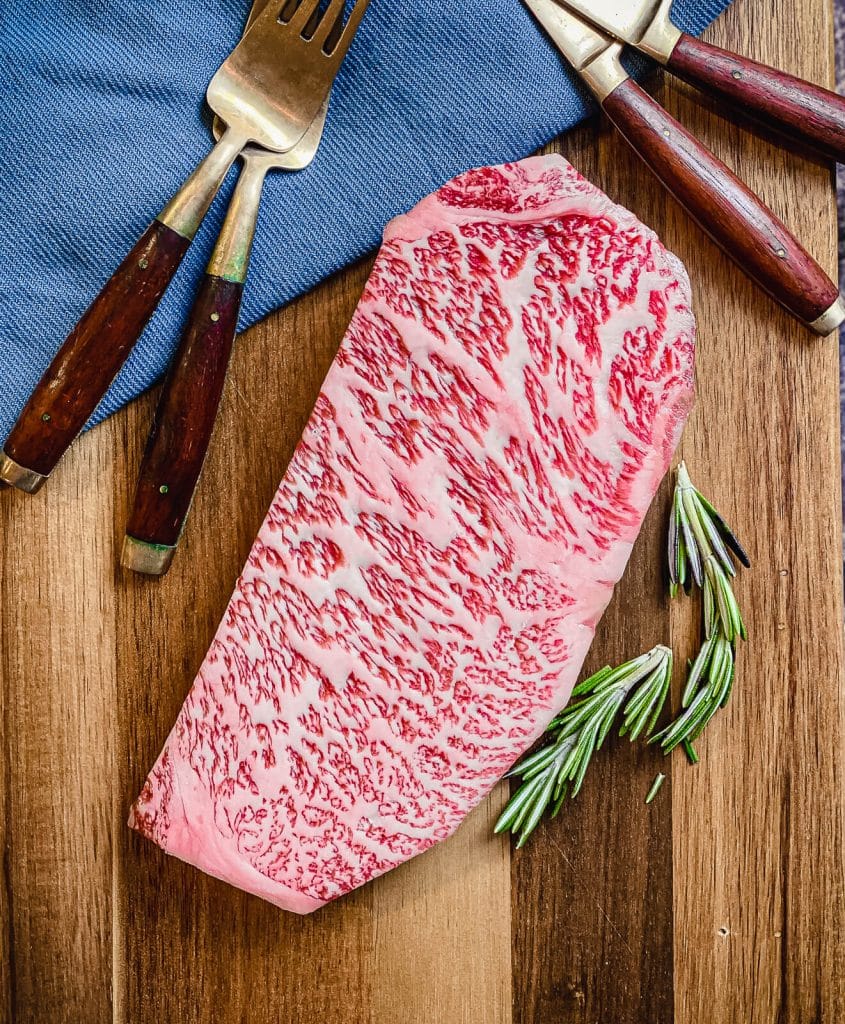 uncooked wagyu strip steak on a cutting board with rosemary