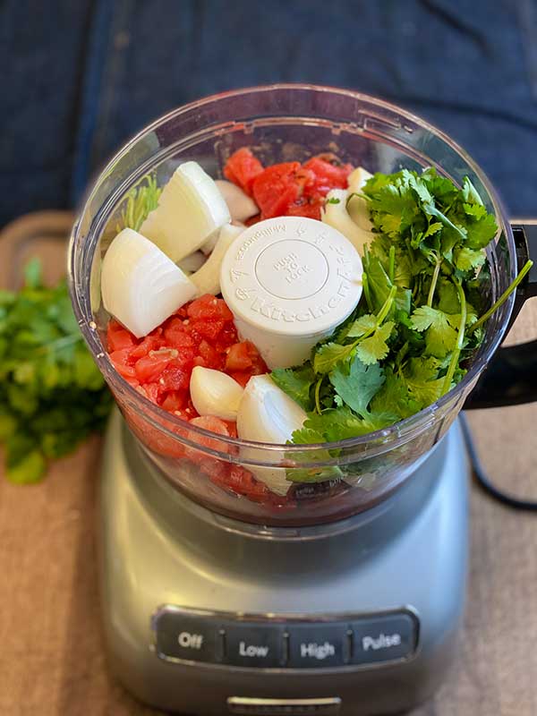 Ingredients for the restaurant style salsa in a food processor ready to be processed: chicken bouillon, garlic, onion, lime, cilantro, salt, and canned tomatoes.