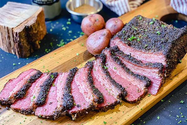 Beer-Braised-Corned-Beef-Feature-Image Smoked and beer braised corned beef on a cutting board with potatoes and a block of hickory wood in the background