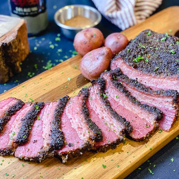 A cutting board with slices of corned beef and potatoes in front of a block of hickory wood and spices