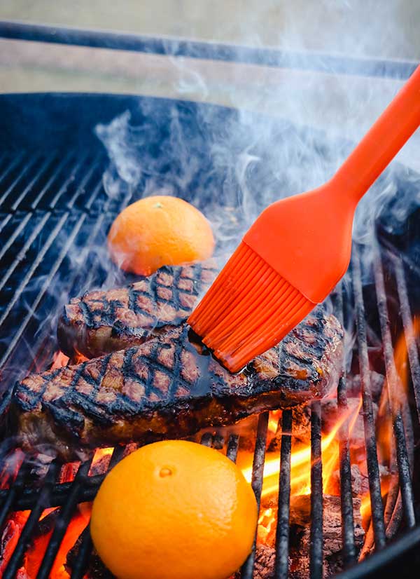 Basting strip steaks on a grill with orange halves