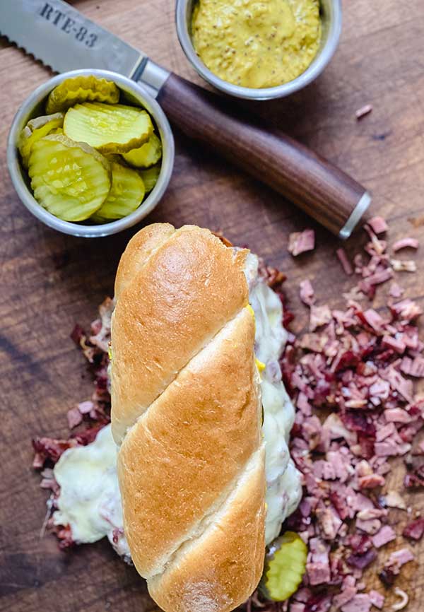 French dip pastrami sandwich on a cutting board, overhead shot with pickles and mustard in small metal serving bowls at top