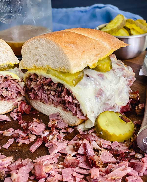close up of French dip pastrami sandwich on a cutting board sliced in half in front of au jus and pickles