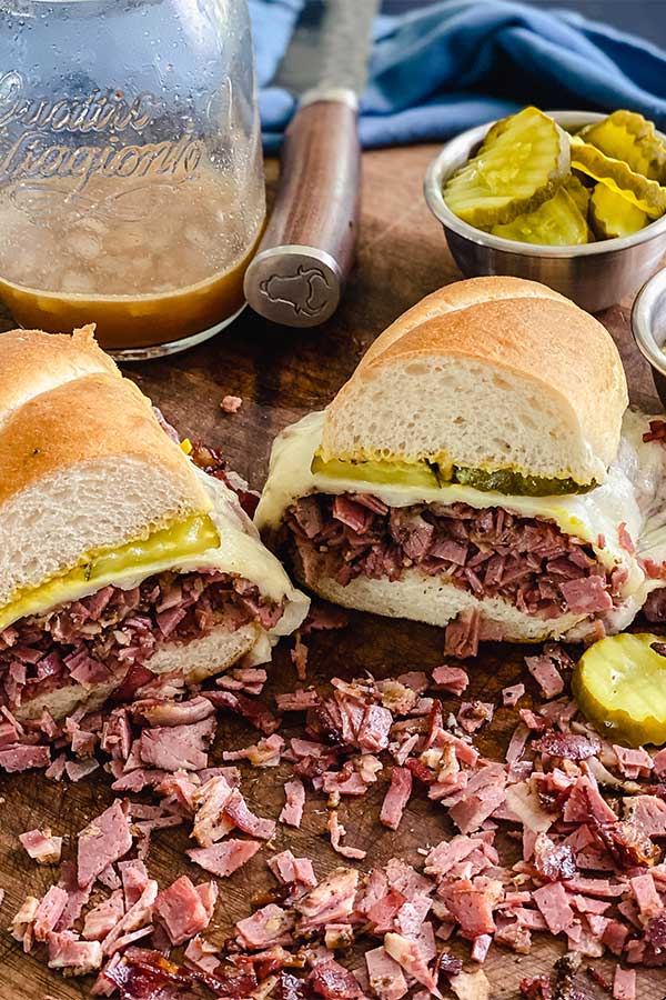 french dip pastrami sandwich cut in half in front of au jus, a knife, and a small bowl of pickles