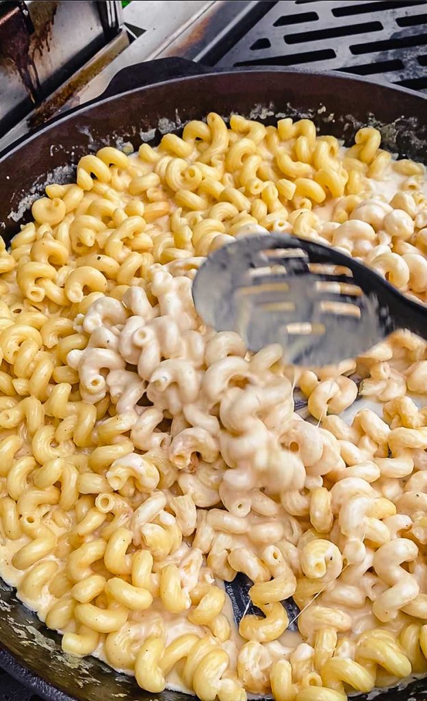 stirring pasta and cheese sauce in cast iron skillet on grill