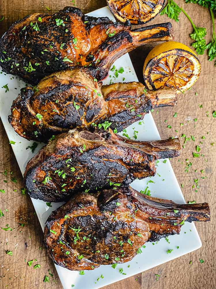 grilled veal chops sprinkled with parsley on a rectangular white plate next to grilled lemons