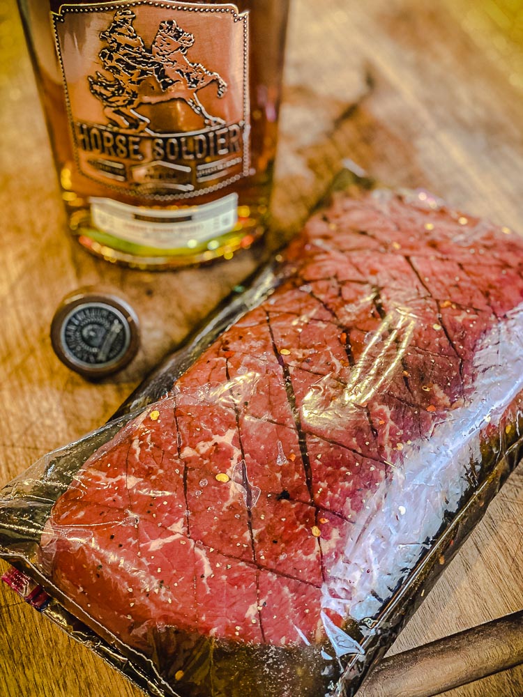 london broil in plastic bag with bourbon marinade and bourbon bottle in background.