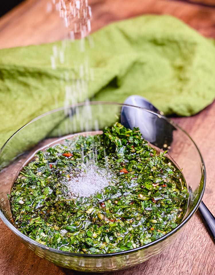 salt being added to a bowl of chimichurri sauce 