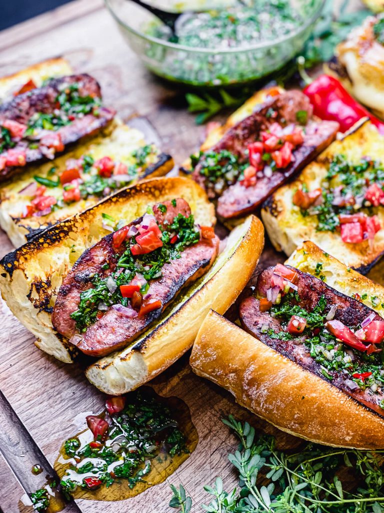 Choripan sitting on a wooden cutting board with fresh herbs and chimichurri sauce