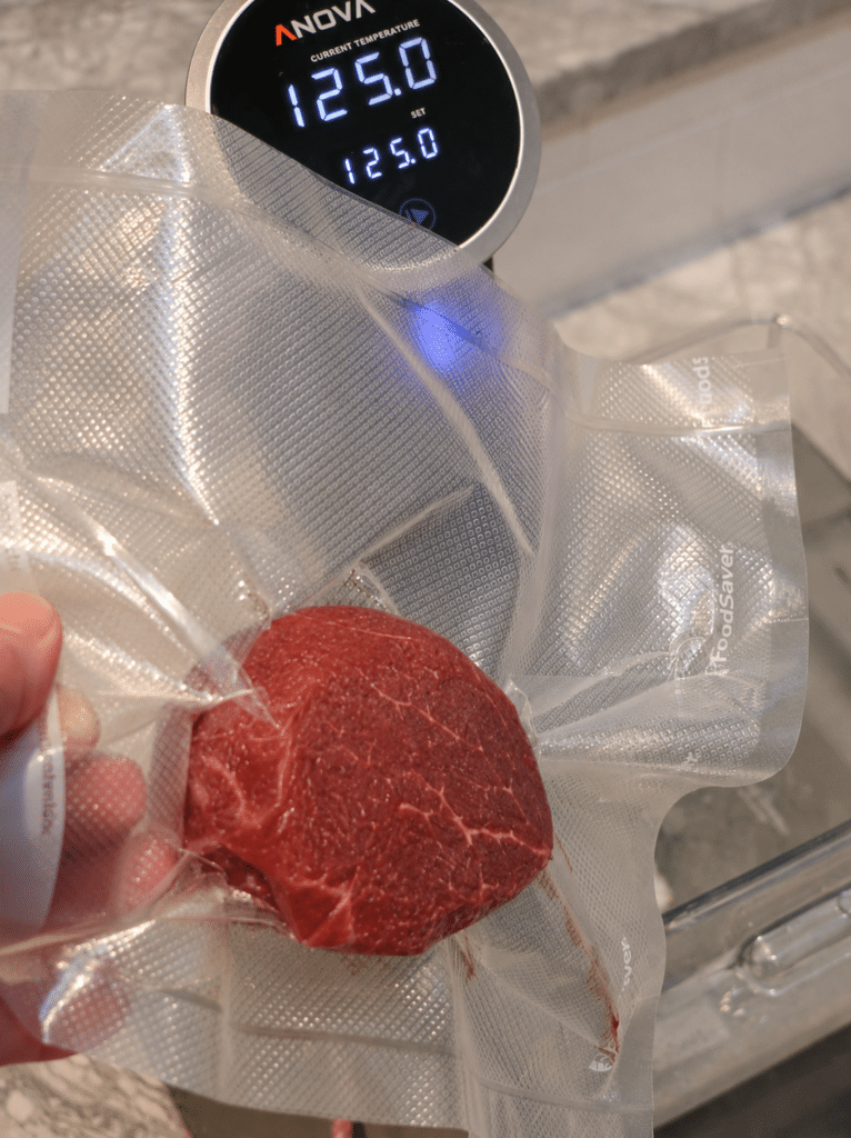 Hand holding a vacuum-sealed filet mignon in front of a sous vide circulator reading 125 degrees before lowering it into the water bath