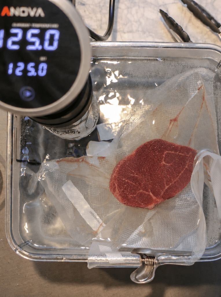 Filet mignon in a vacuum-sealed bag clipped inside a sous vide water bath with an Anova circulator set to 125 degrees for medium rare