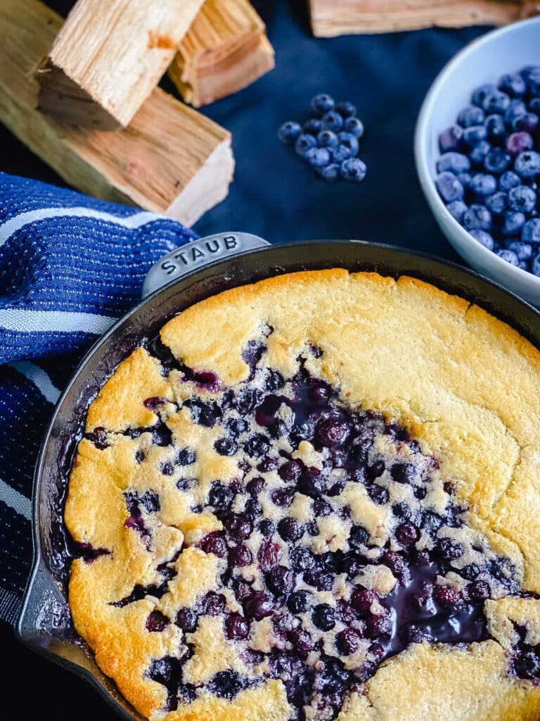 smoked blueberry cobbler in a cast iron pan with a bowl of blueberries setting next to it