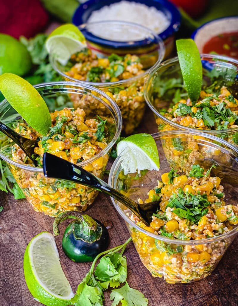 cups of esquites sitting on a cutting board along with some limes and cilantro