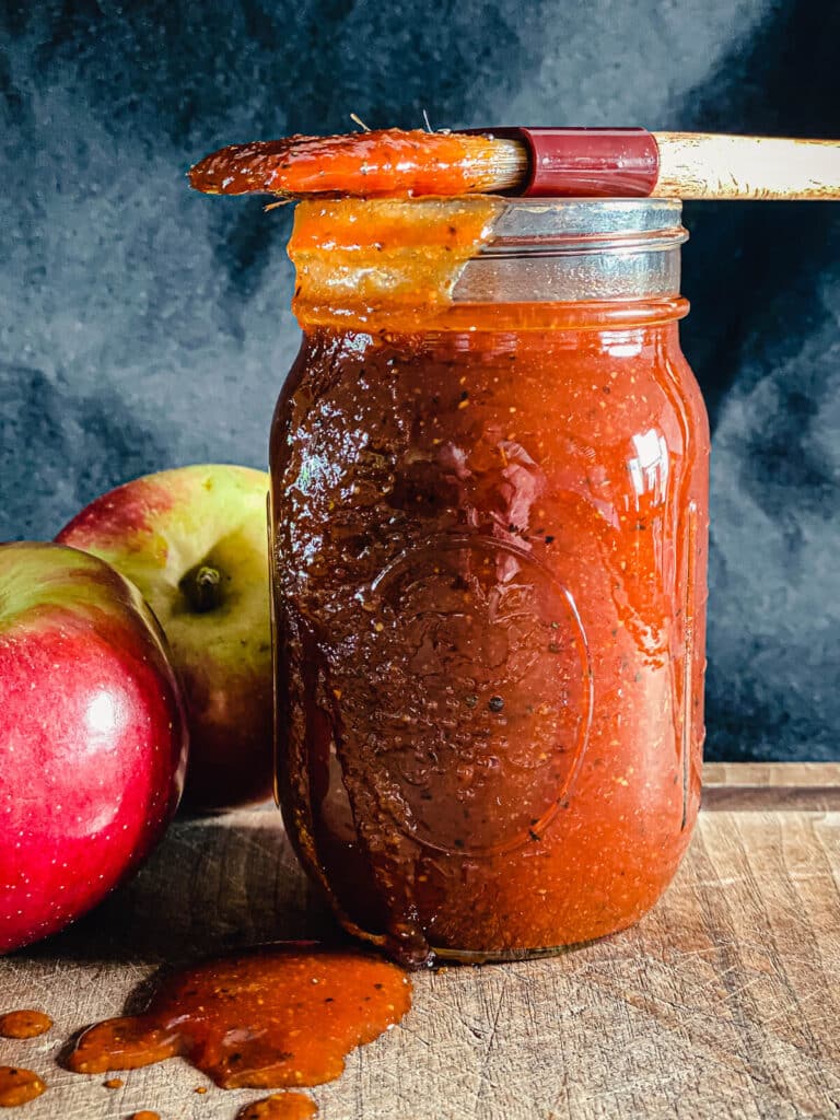 Jar of apple whiskey BBQ sauce with a brush on top and apples in the background