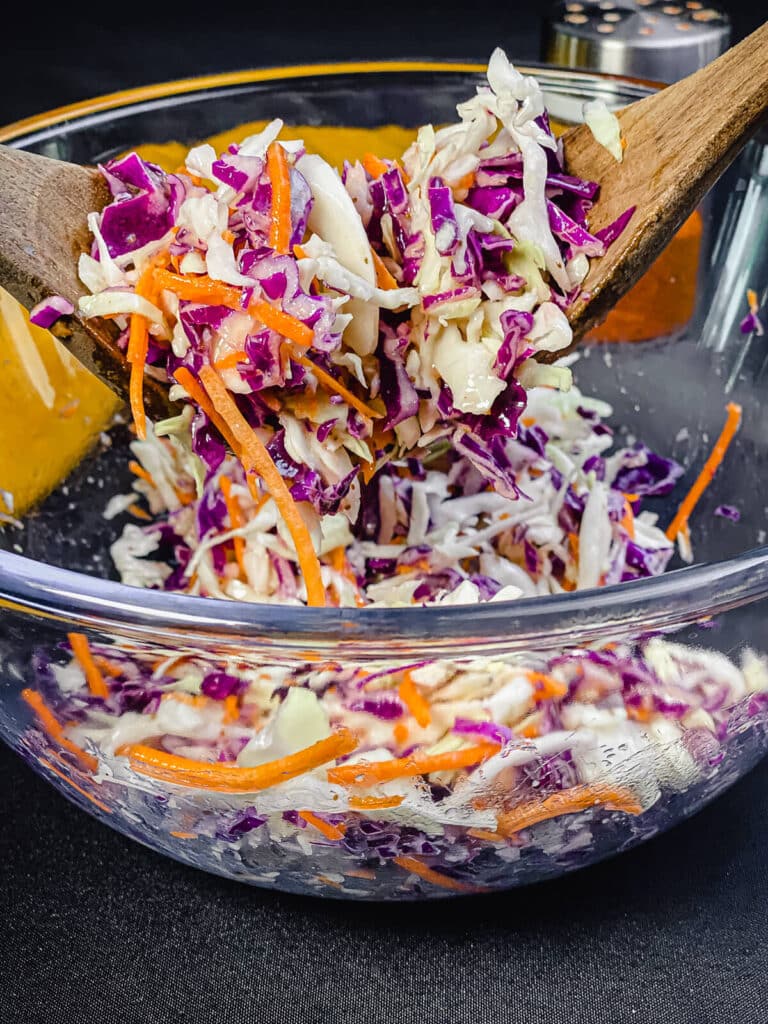 Salad tongs tossing a no mayo coleslaw mixture in a large glass bowl