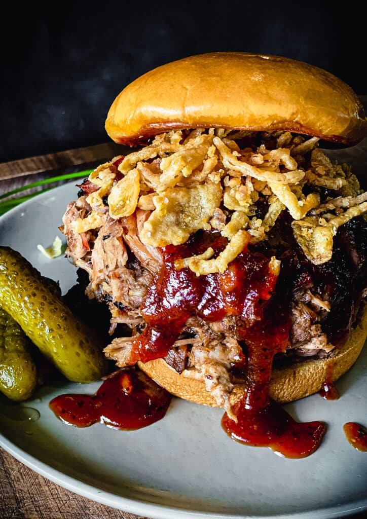 apple brined pulled pork sandwich on a bun with bbq sauce dripping down and pickles on the plate