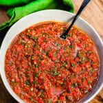 a bowl of red chimichurri on a cutting board with a black spoon and green towel