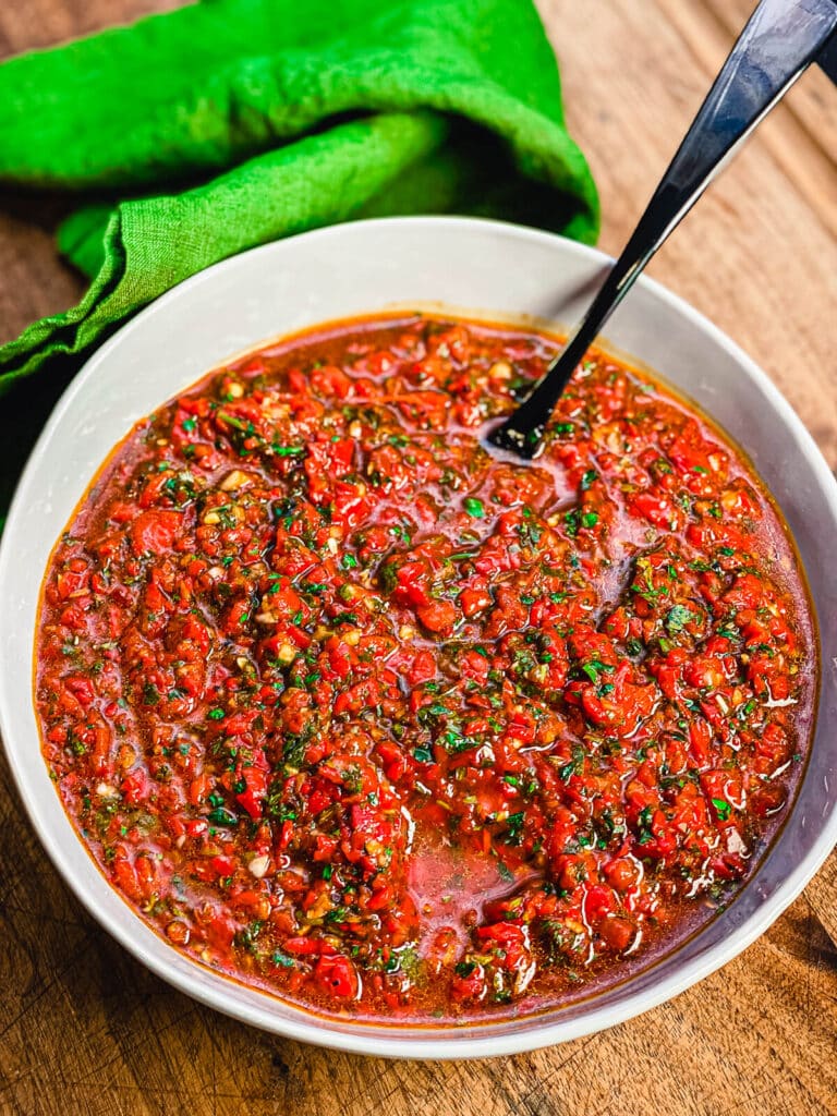 a bowl of red chimichurri on a cutting board with a black spoon and green towel