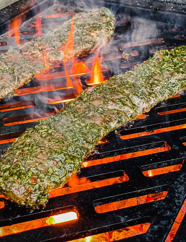 chimichurri covered skirt steaks on a grill over hot coals with flames