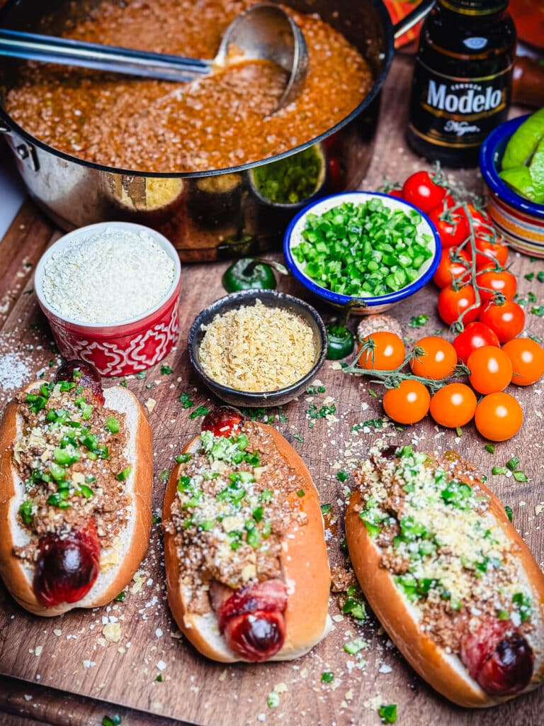 taco dogs on a cutting board with chili, toppings, and beer in the bckground