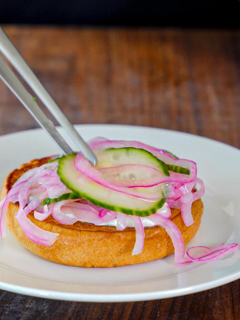 cucumbers and red onions being added to a bun