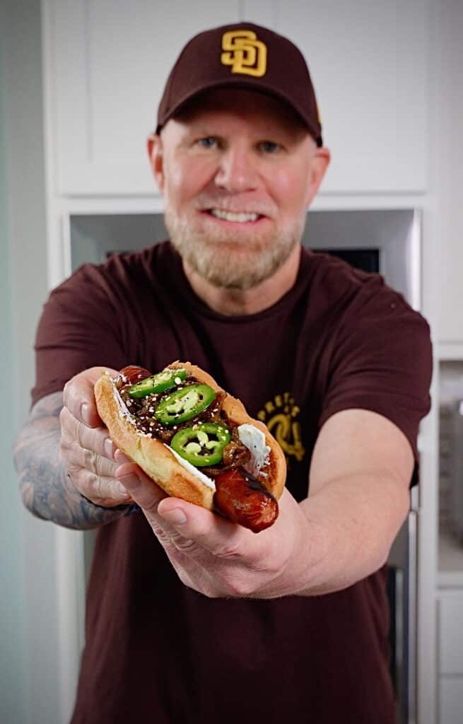 man in hat holding a seattle style hot dog with jalapenos